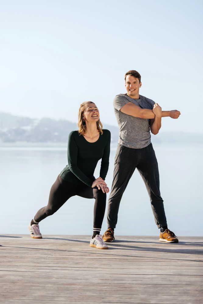 Sportliches Paar dehnt sich gemeinsam am Seeufer bei Sonnenaufgang – Sanopal Basic unterstützt Energie und Vitalität vor körperlicher Aktivität