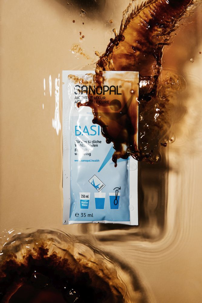 Sanopal Basic Sachet mit spritzender dunkler Trinklösung, close-up – Aktive Zellkur mit fruchtigem Geschmack zum Auflösen in Wasser