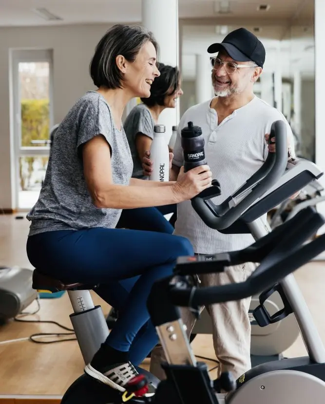 Lächelndes Paar im Fitnessstudio – Frau auf Fahrradergometer hält Sanopal Trinkflasche, Mann mit Cap im Gespräch – Healthy Aging und aktiver Lebensstil mit Sanopal Basic im fortgeschrittenen Alter