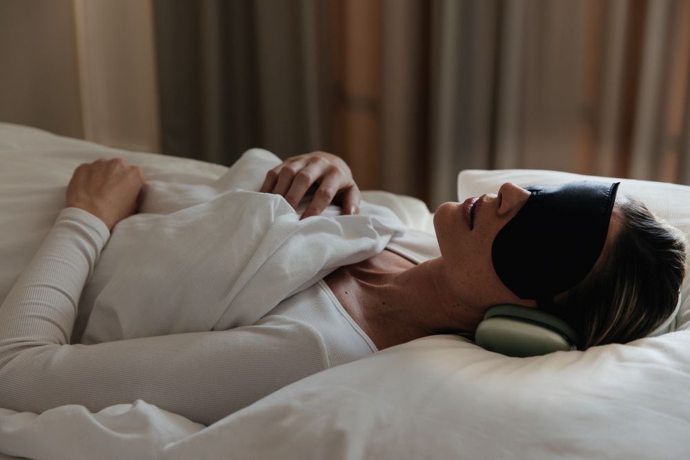Frau mit Schlafmaske und Kopfhörern liegt erschöpft im Bett – Sanopal Forte Wirkstoffkombination A5H mit Alpha-Ketoglutarat und 5-Hydroxymethylfurfural unterstützt Zellen und Mitochondrien bei Long COVID, Fatigue und posttraumatischem Syndrom